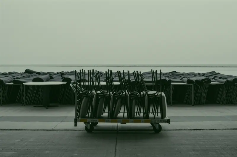 Stacked event chairs, folded round tables and a rolled carpet on a loading apron behind a building.