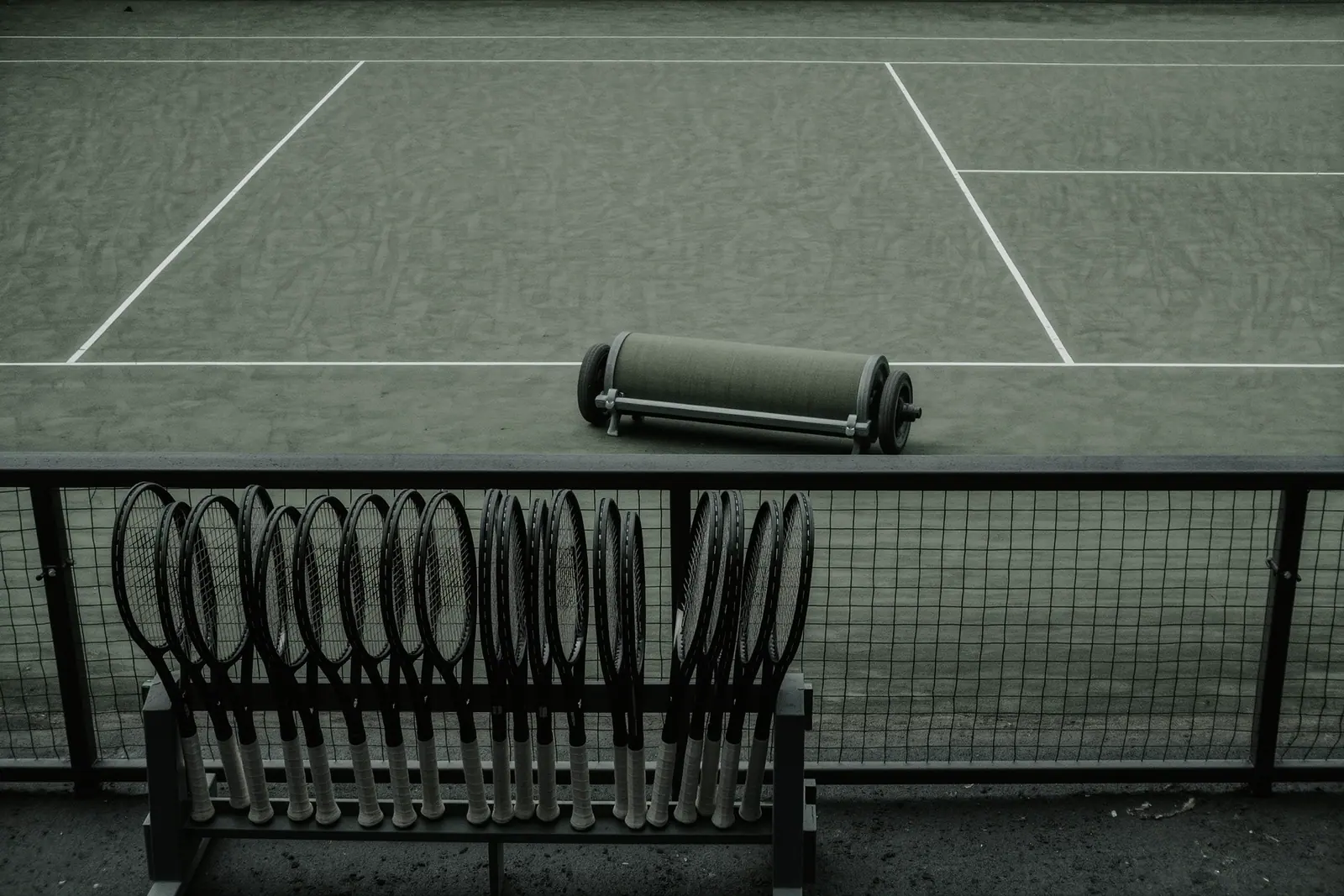 An empty clay tennis court behind a wire fence with a heavy net roller parked at the baseline, a rack of hire rackets standing beside the fence in the foreground.