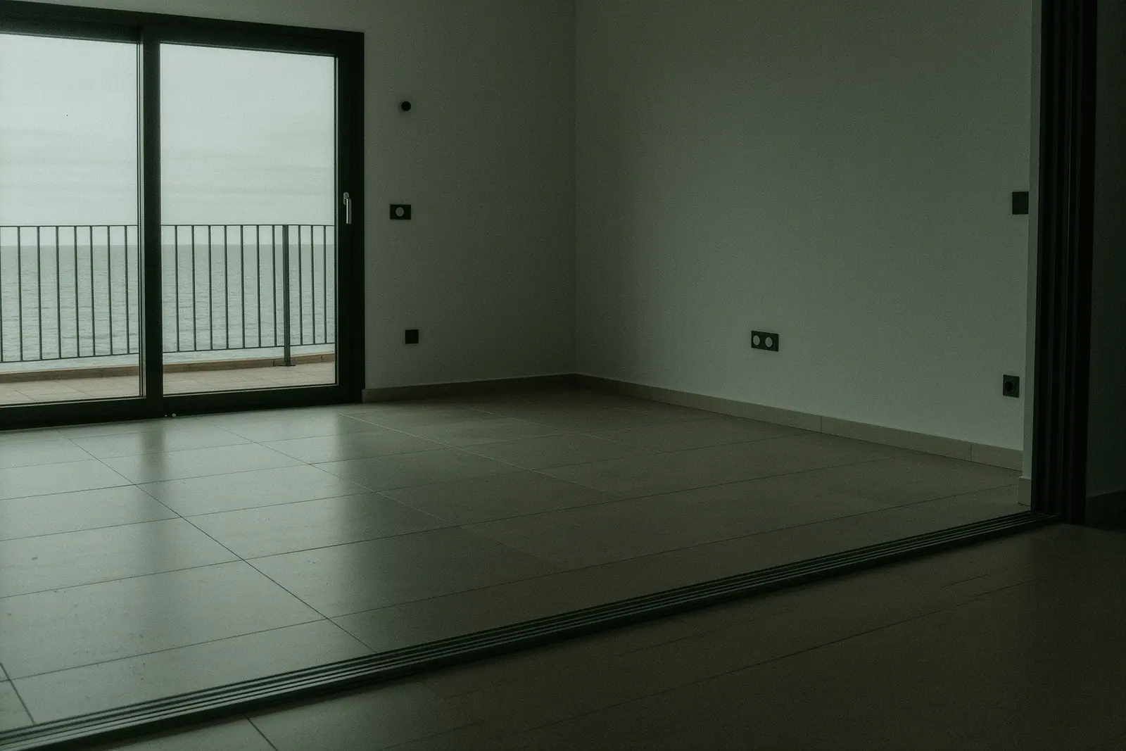 An empty modern apartment living room with sliding doors onto a balcony and bare walls, the tiled floor and door track running across the foreground, flat grey sea beyond.