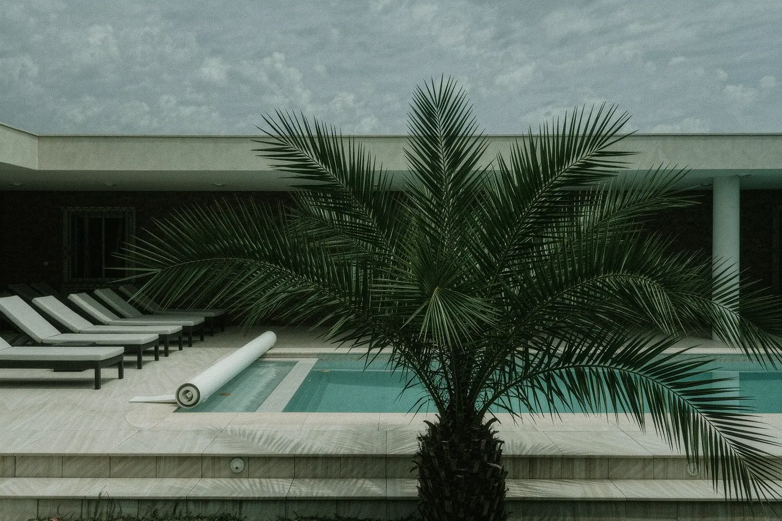 An empty villa pool terrace with sun loungers stacked to one side and the pool cover half rolled back, a palm leaning over the pale tiles in the foreground.
