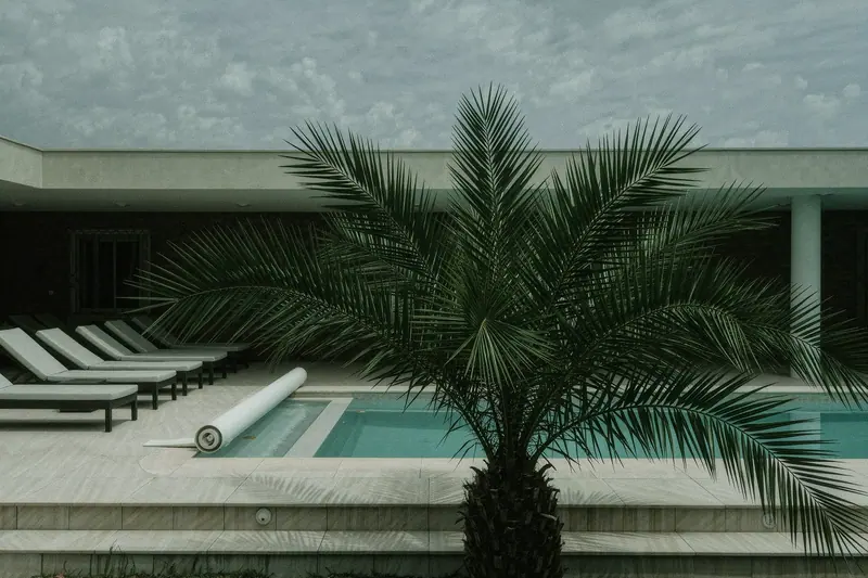 An empty villa pool terrace, sun loungers stacked to one side, the pool cover half rolled and palm shade falling across the tiles.