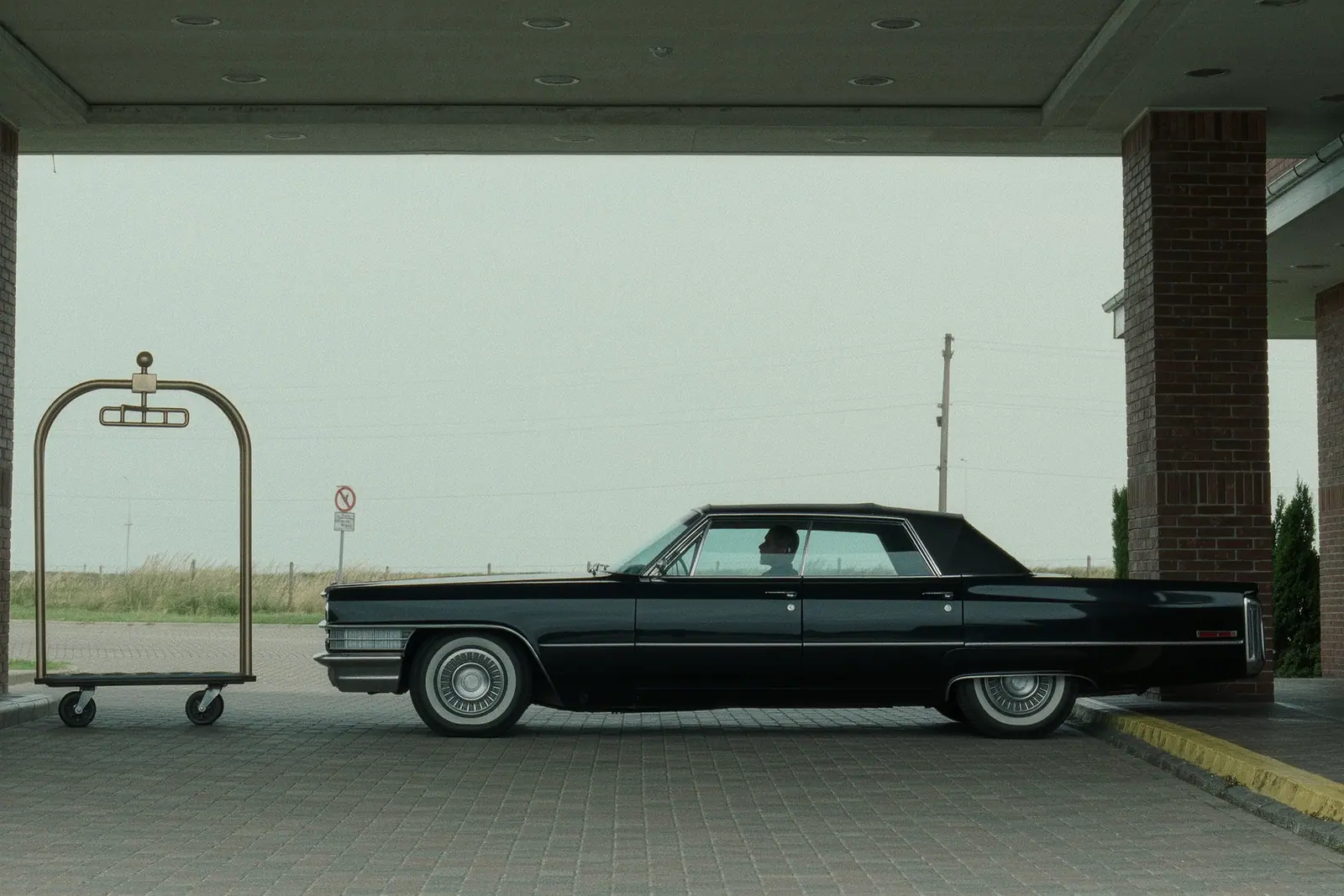 A black saloon waiting under a hotel entrance canopy with its driver's door shut and nobody around, an empty luggage trolley standing to one side on the paved drive.