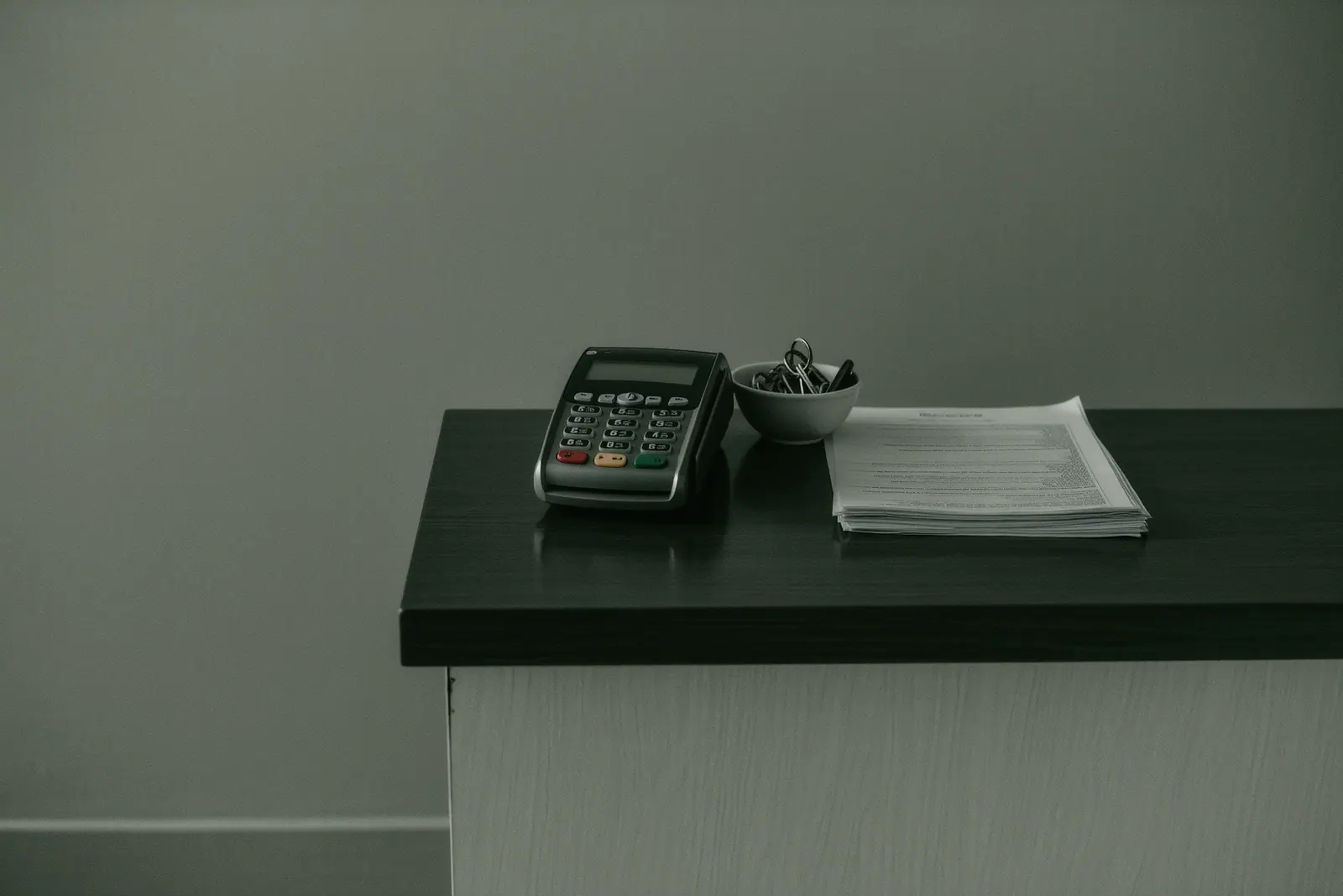 A plain rental desk counter with a card terminal, a stack of paper contracts and a bowl of key fobs, the corner of the counter and the terminal close in the foreground.