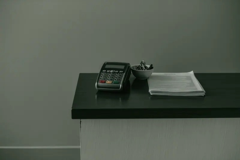 A plain rental desk counter with a card terminal, a stack of printed contracts and a bowl of key fobs.