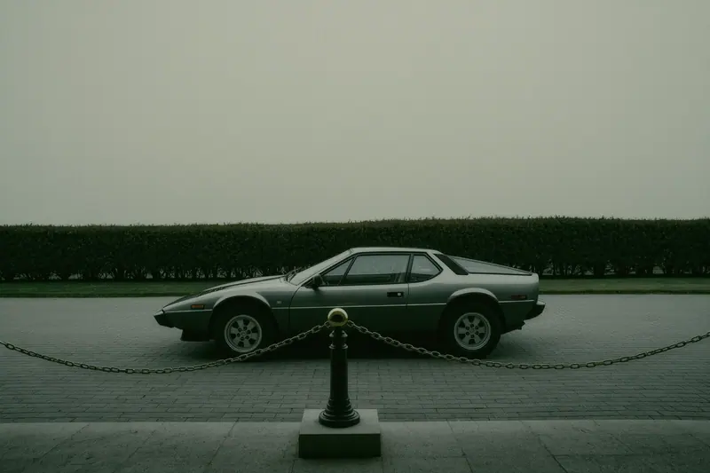 A low unbranded sports car parked on a plain paved forecourt behind a chain barrier, a clipped hedge and blank wall behind it.