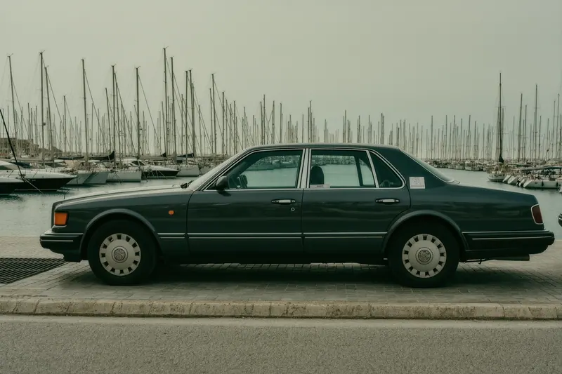 A dark unbranded saloon stopped at a marina kerb in Puerto Banús with a valet ticket tucked under the wiper, masts of moored boats behind.