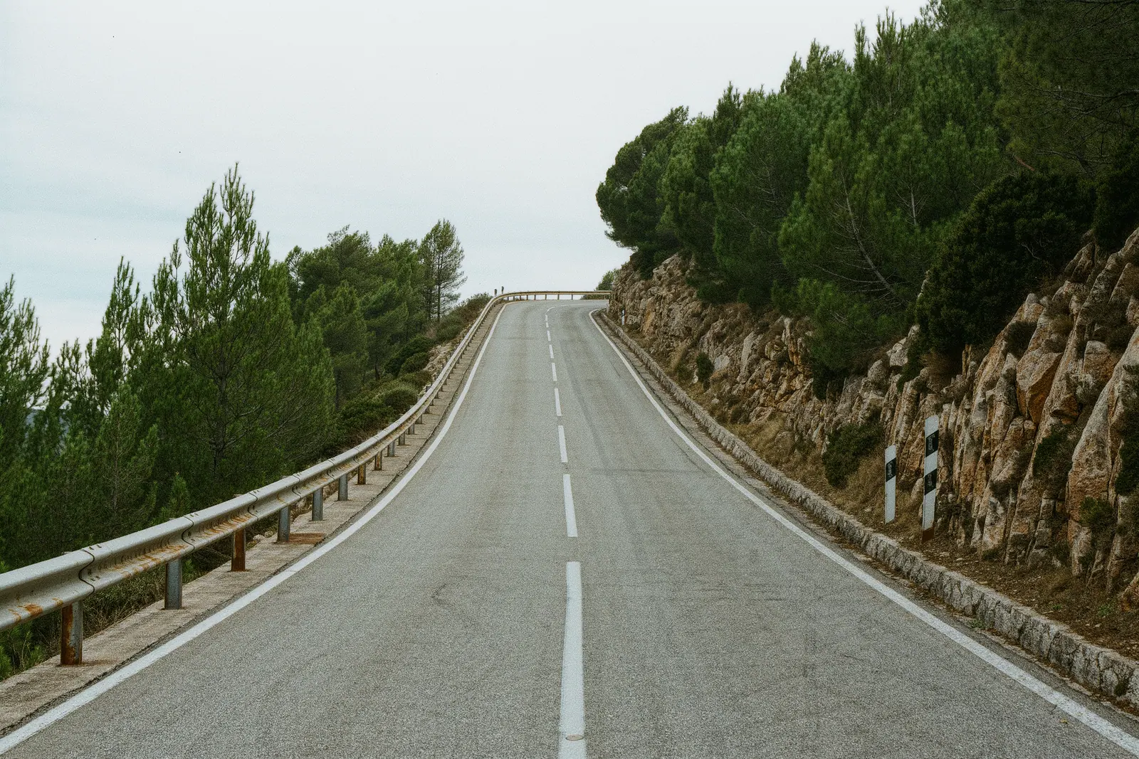 An empty mountain road above Marbella climbing into pine and bare rock, a weathered steel crash barrier running along the outside edge in the foreground.