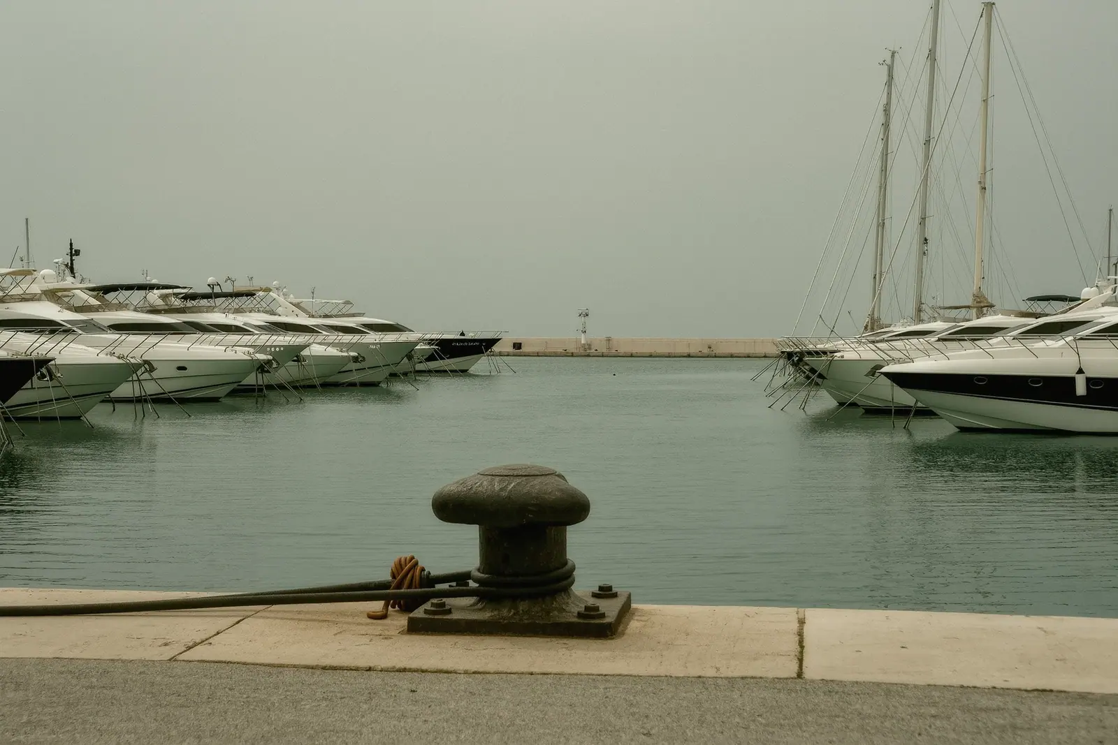 Motor yachts lying stern-to along the quay at Puerto Banus with their passerelles down, a heavy iron bollard and a coiled water hose on the concrete in the foreground.