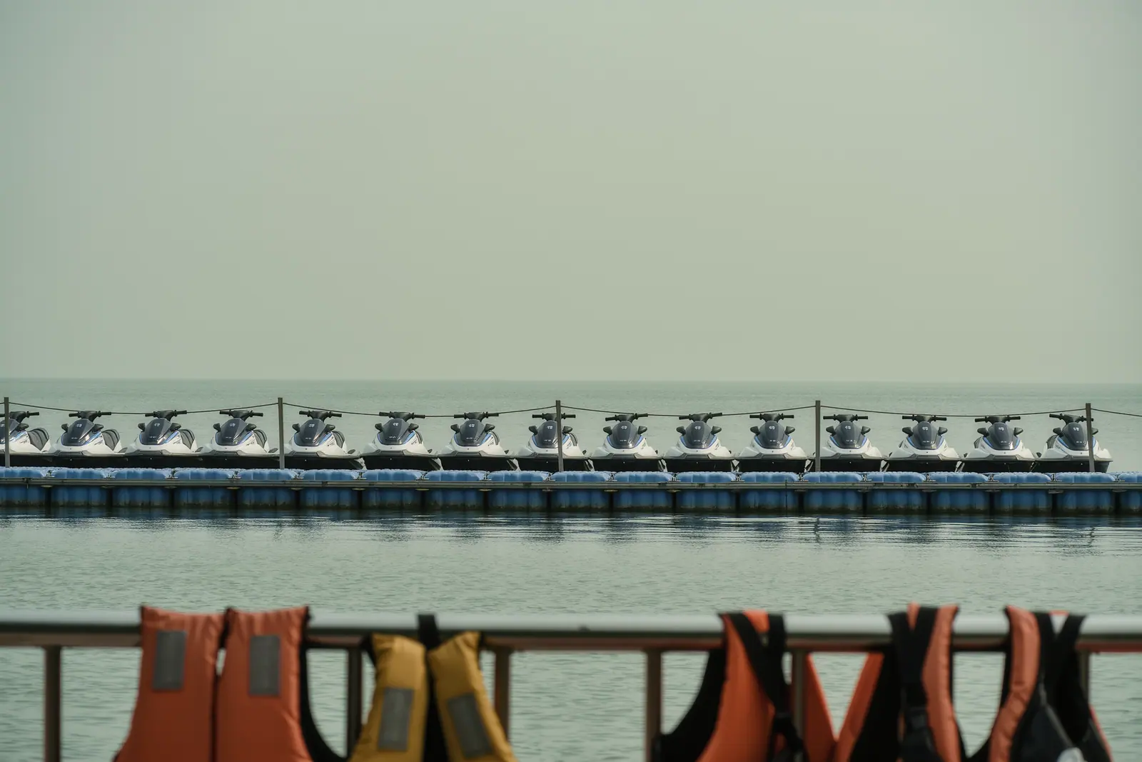 Jet skis lined up in a row on a floating pontoon behind a slack rope barrier, life jackets hanging over a metal rail in the foreground.
